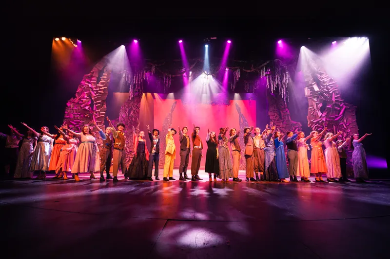 A full cast on a colorfully lit theater stage